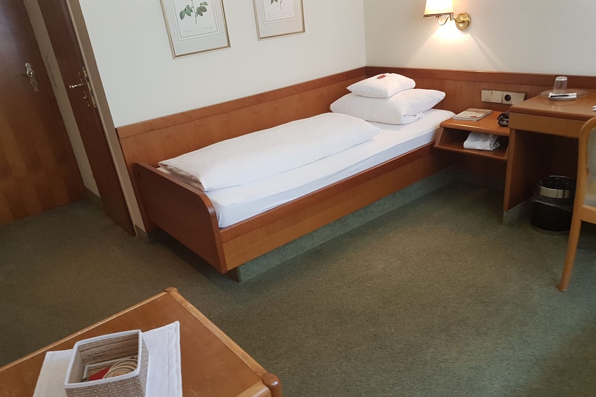 Uma cama de solteiro com roupa de cama branca, cabeceira de madeira, mesa de cabeceira com abajur e uma prateleira pequena com um cesto de lixo.