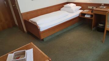 A single bed with white linens, a wooden headboard, a bedside table with a lamp, and a small shelf with a trash bin.
