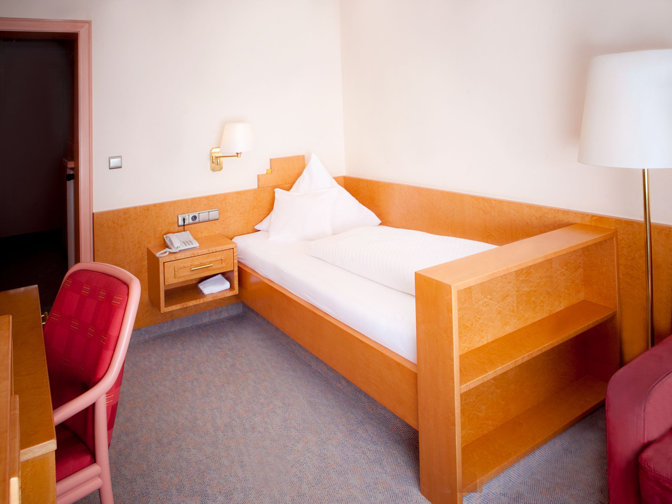 Une chambre d’hôtel avec un lit simple, une table de chevet avec une lampe, une chaise et un bureau.