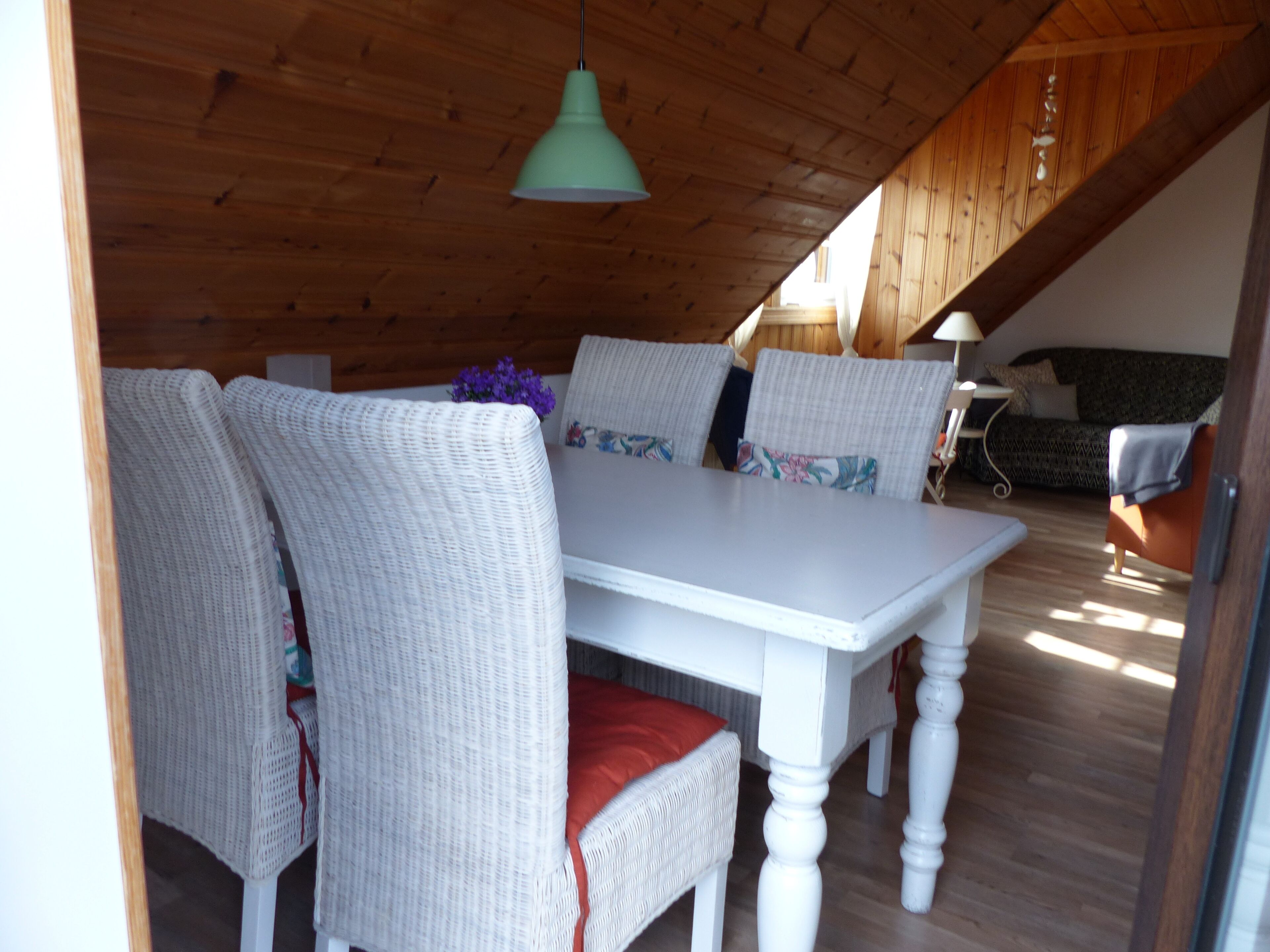 Uma sala de jantar com uma mesa branca e cadeiras de vime, um lustre pendente e teto de madeira.