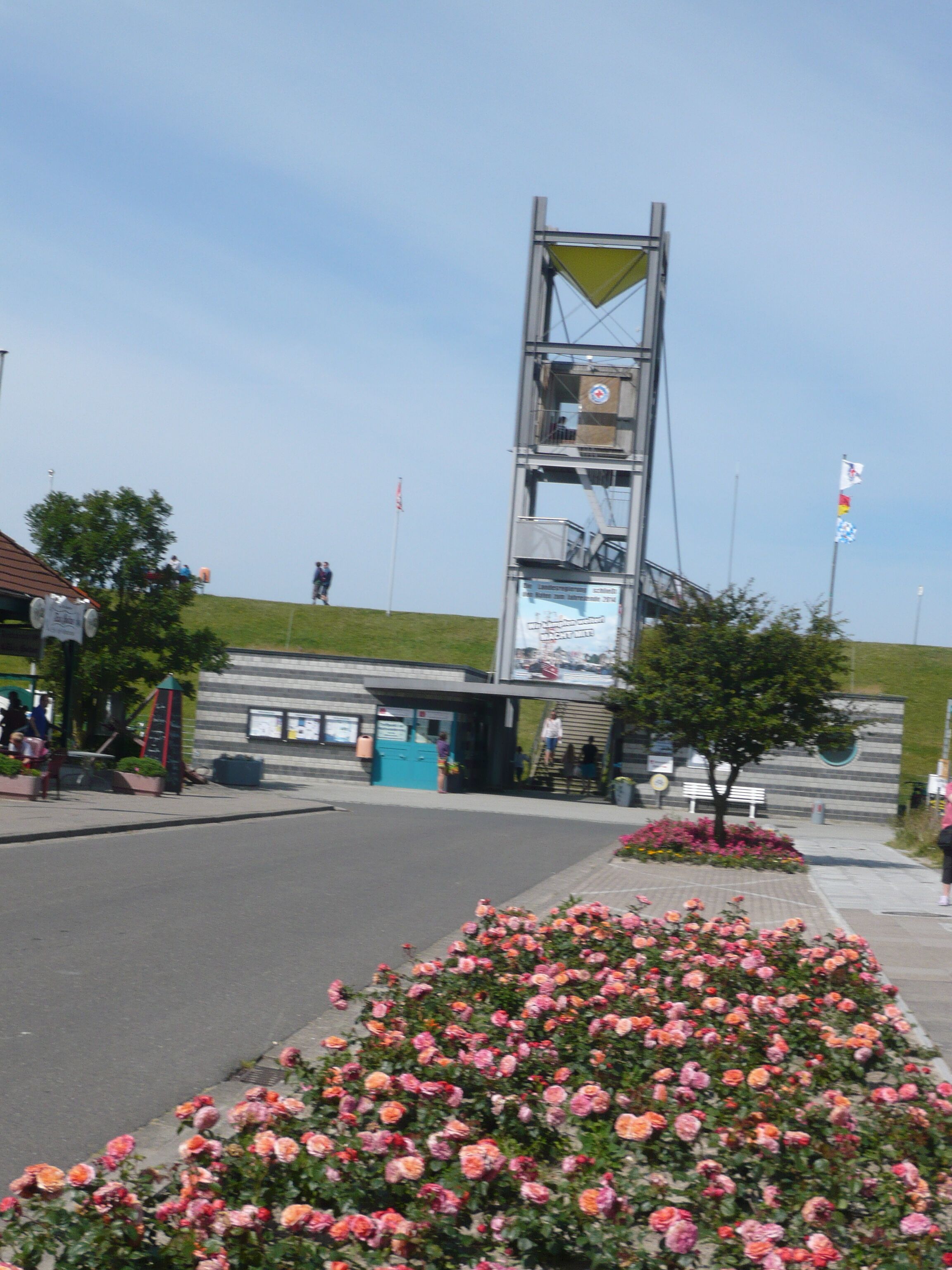 A public square with a tall, modern structure, a flower bed with pink flowers, and a grassy hill in the background.