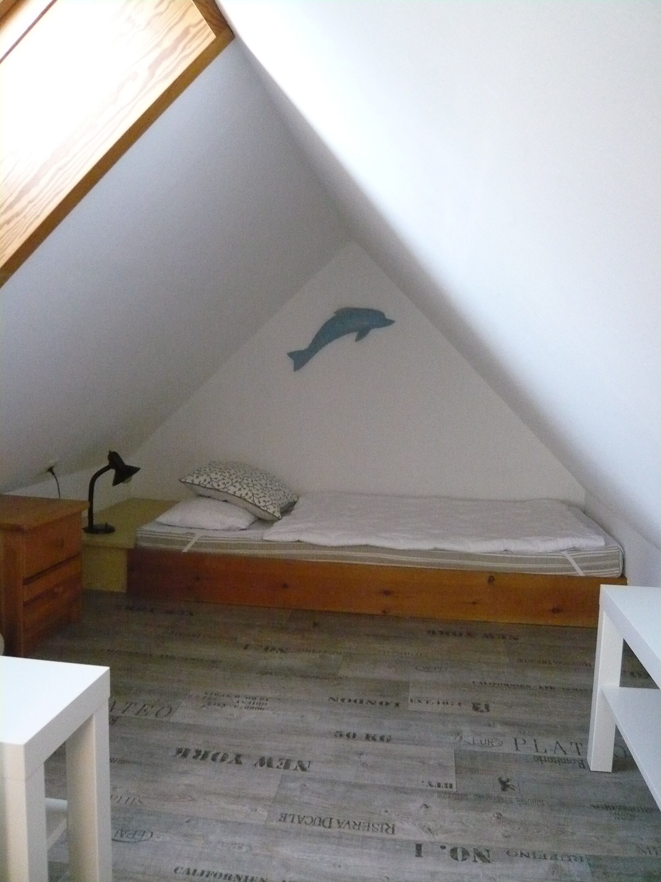 Een kleine, schuine kamer met een enkel bed, een houten nachtkastje, een lamp en een sticker van een dolfijn op de muur.