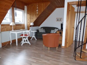 A loft-style room with a sloped ceiling, wooden walls, a dining table with chairs, a sofa, and a staircase.
