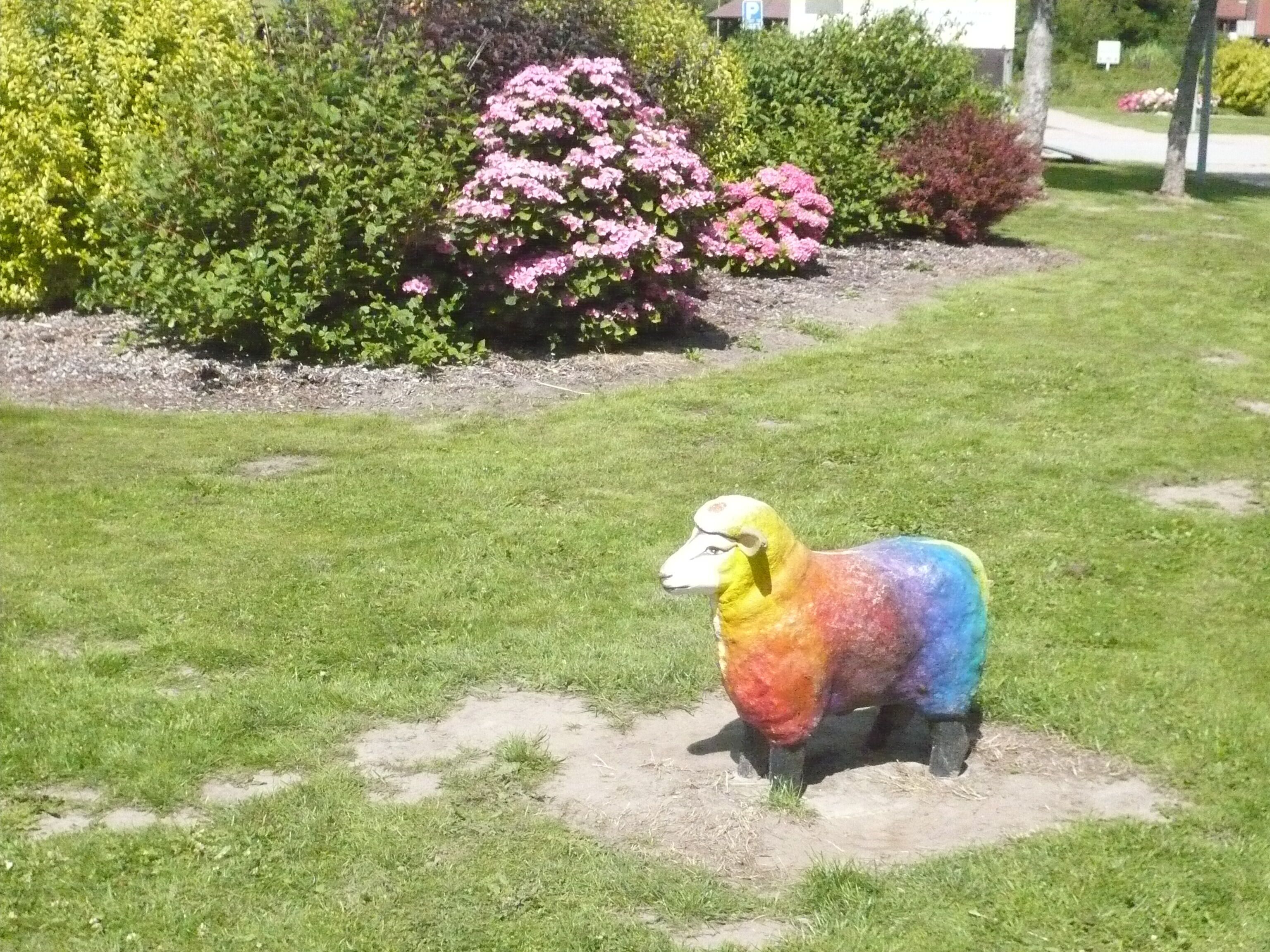 Eine farbenfrohe Schafskulptur in einer grünen Landschaft, im Hintergrund verschiedene Pflanzen und Sträucher.