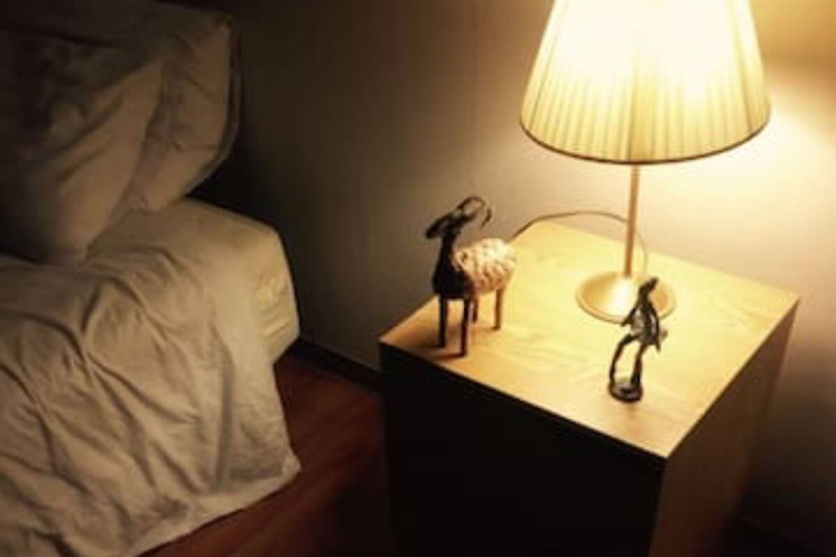 A small bedside table with a lamp and two figurines, a bed with a beige comforter, and a wooden floor.