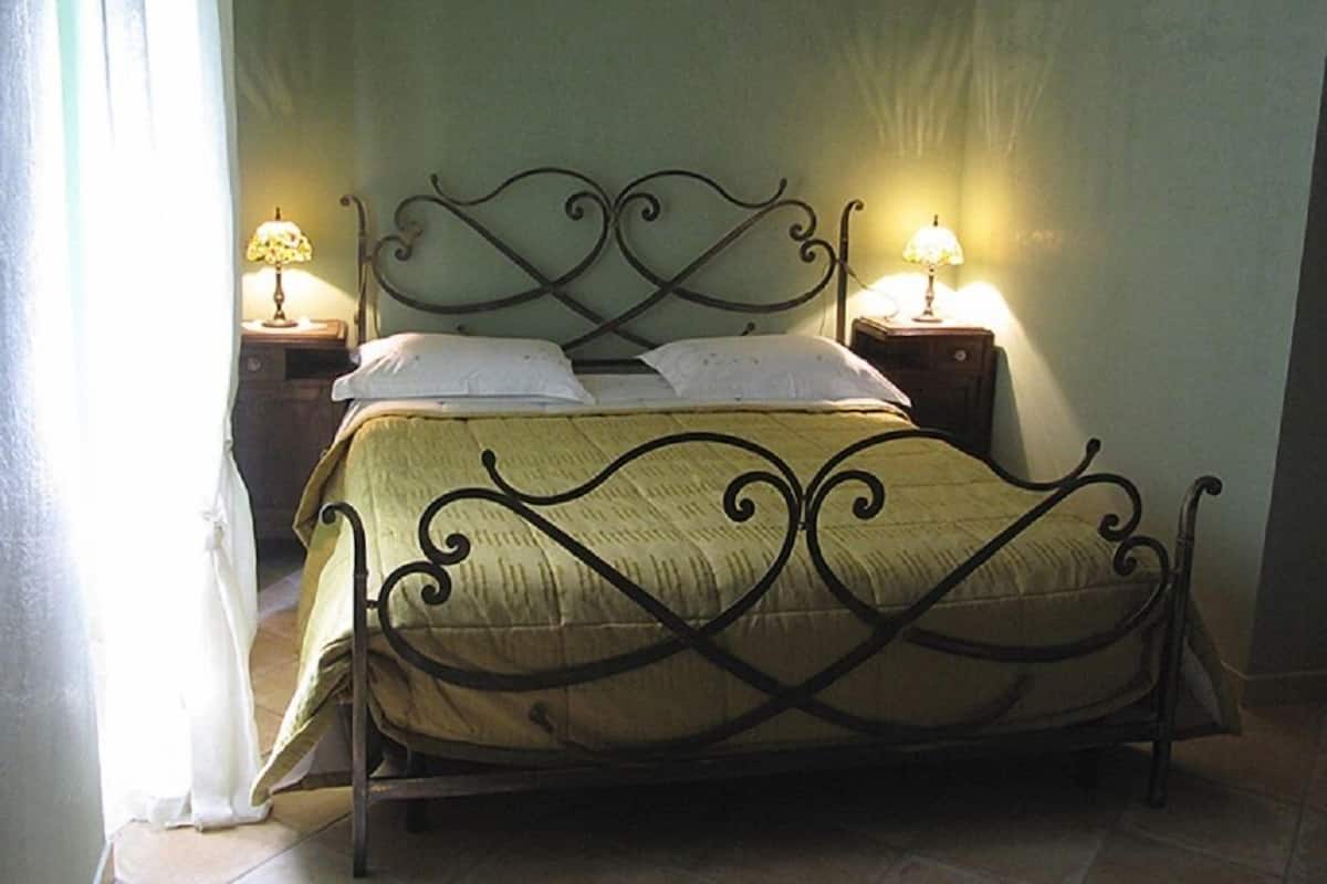 A bed with a metal frame and a beige bedspread, flanked by two bedside tables with lamps, in a room with plain walls and a tiled floor.
