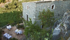 Un edificio de piedra con dos ventanas y una puerta, rodeado de vegetación y un patio con mesas y sillas.