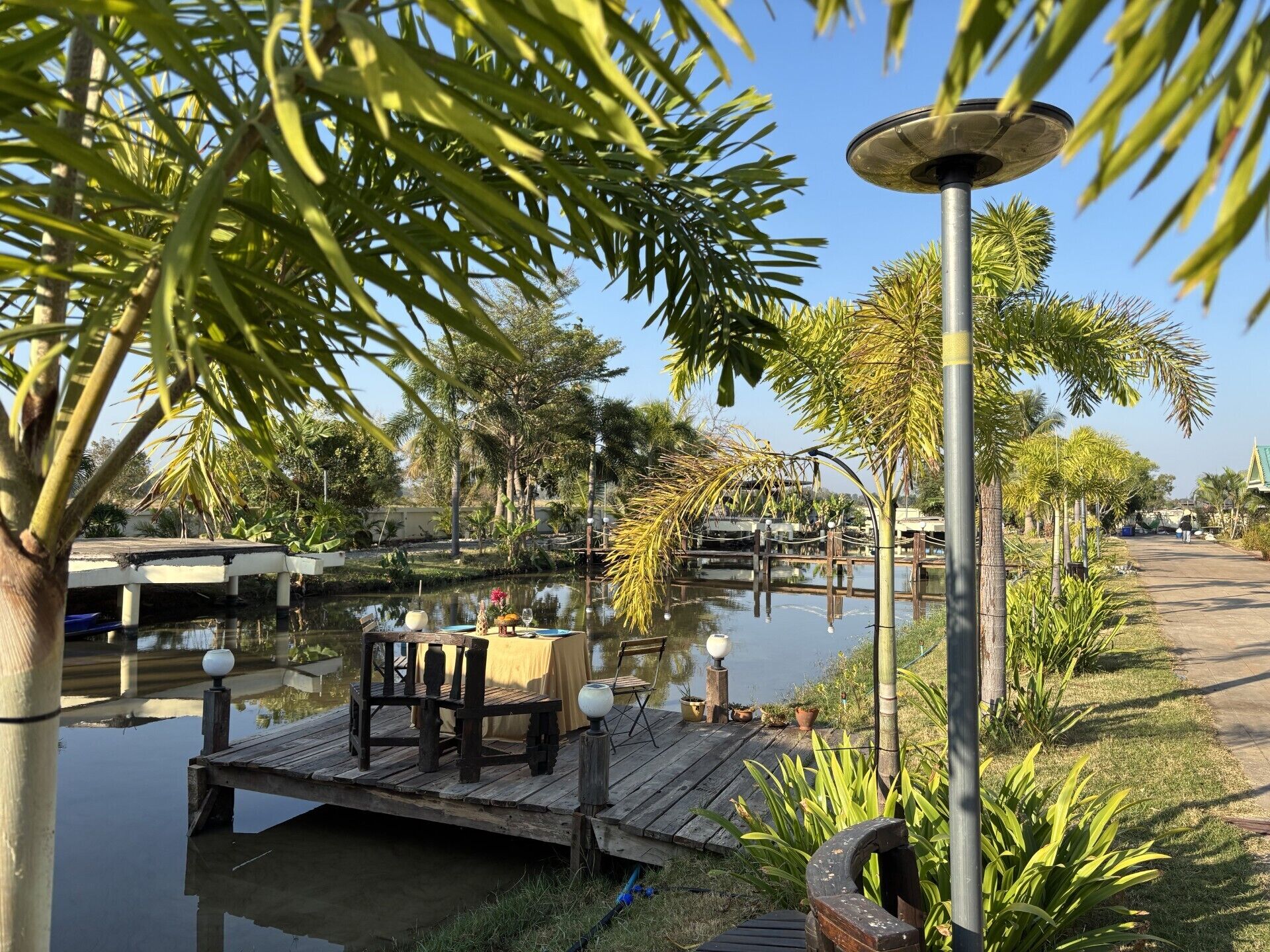 A riverside dining area with a wooden deck, tables, chairs, and a lamp post.