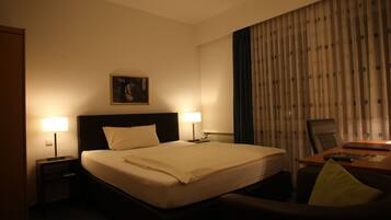 A hotel room with a large bed, two bedside lamps, a desk with a chair, and a window with curtains.