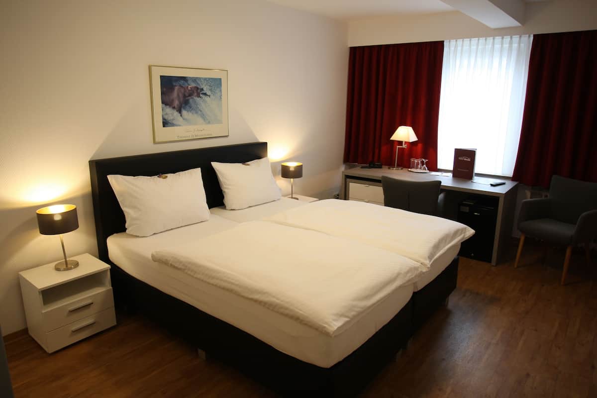 Habitación de hotel con una cama grande, dos mesitas de noche con lámparas, un escritorio con silla y una ventana con cortinas rojas.