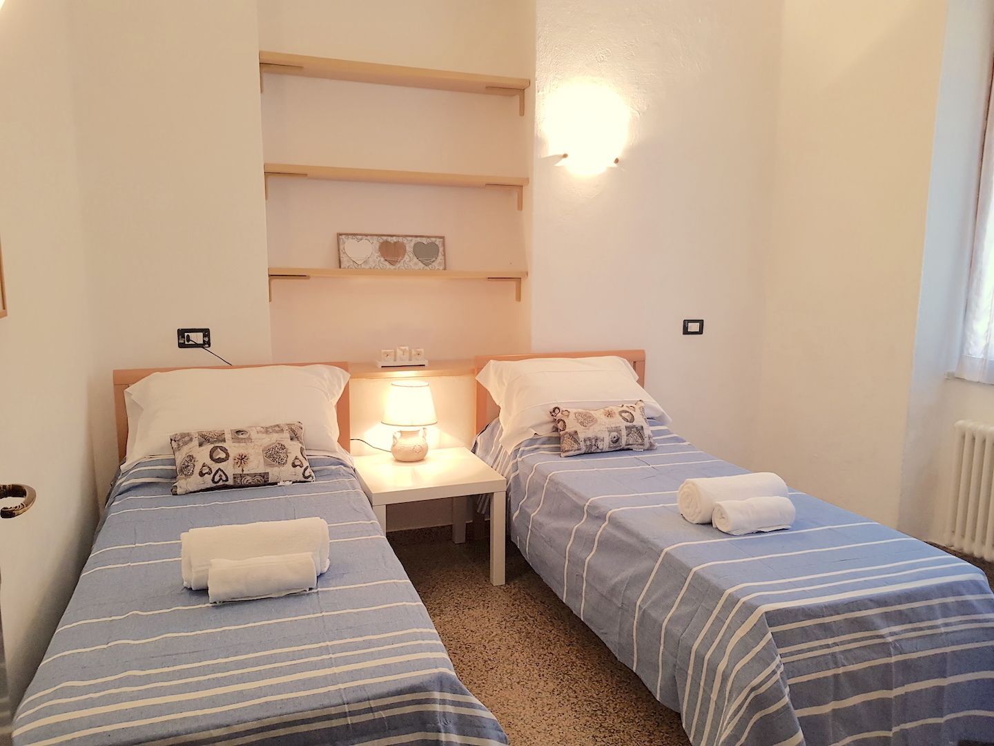 Quarto pequeno e limpo com duas camas individuais, mesa de cabeceira com abajur e prateleira sobre as camas.