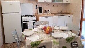 A compact kitchen with a refrigerator, stove, and sink. There is a dining table set with plates, glasses, and a bowl of fruit. - Augusto charming flat with sea view (Monterosso al Mare)