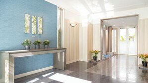 A modern reception area with a blue textured wall, a reception desk, potted plants, and a large window allowing natural light. - Keikyu EX Inn Shinagawa Shimbamba (Tokyo)
