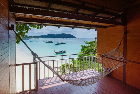 Una hamaca en una terraza de madera con vista a una playa, barcos y una isla distante.