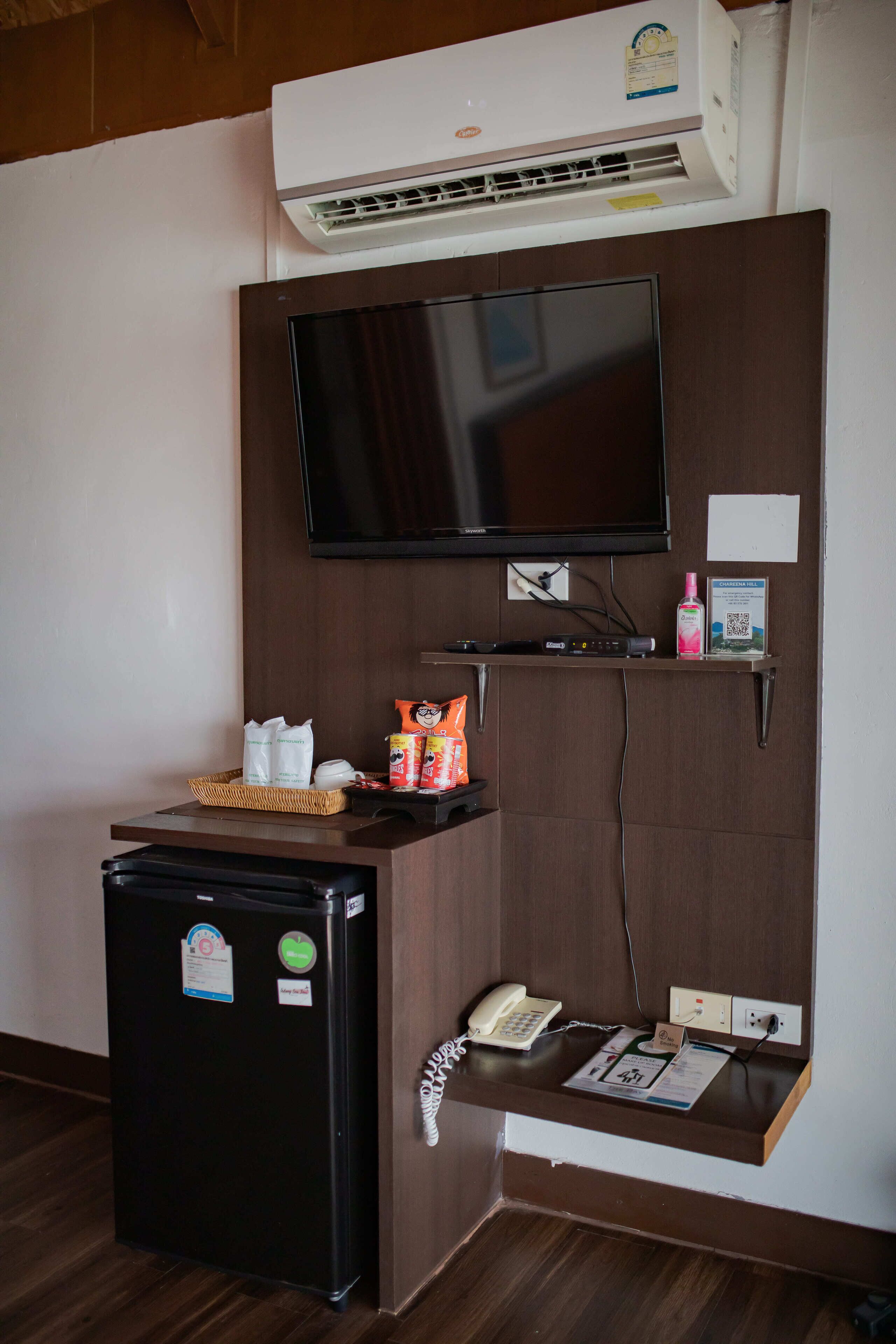A hotel room with a flat-screen TV mounted on the wall, a small refrigerator, a phone, and a shelf with toiletries and a basket.