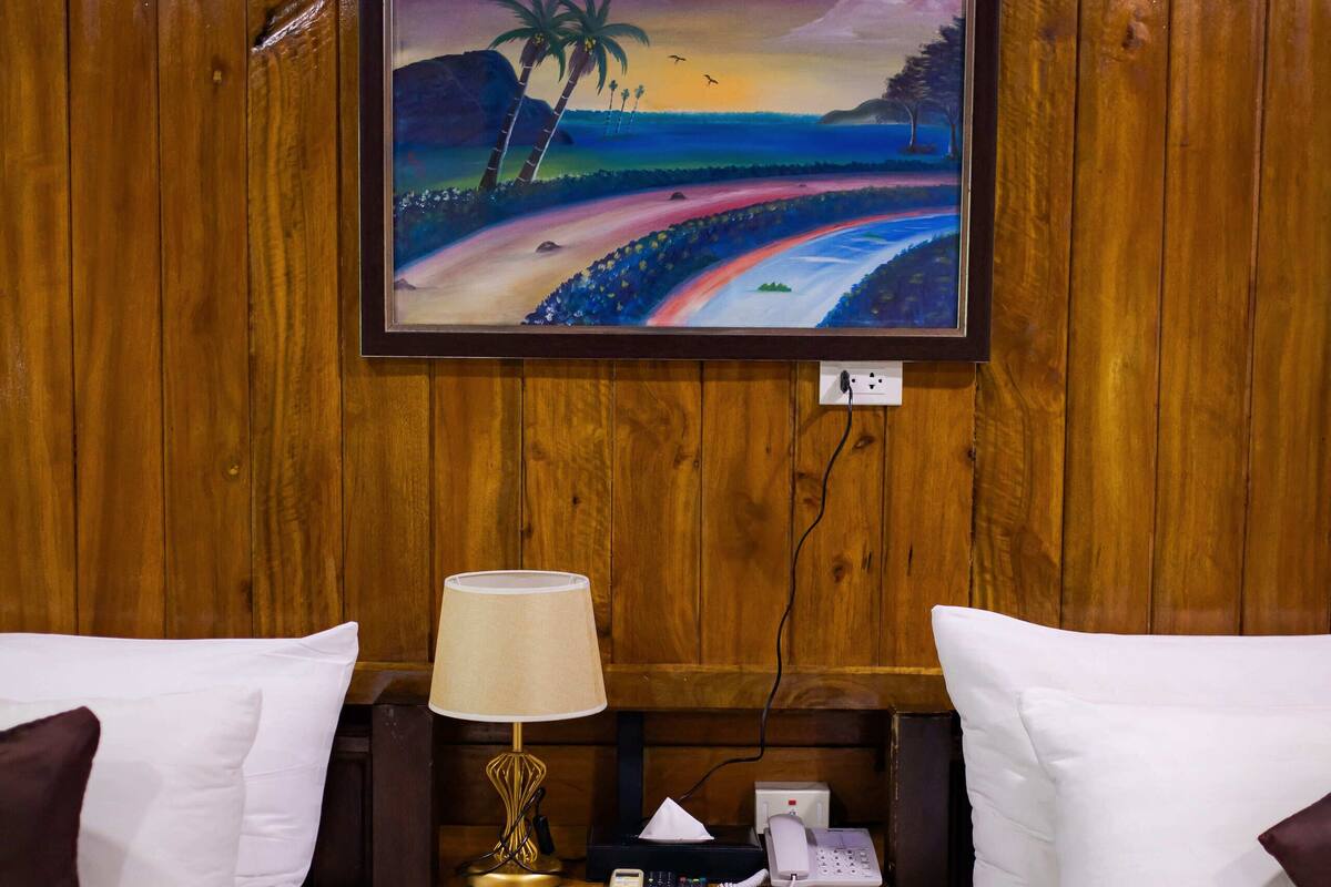 A hotel room with two beds, wooden walls, a painting of a beach scene, a bedside table with a lamp, and a phone.