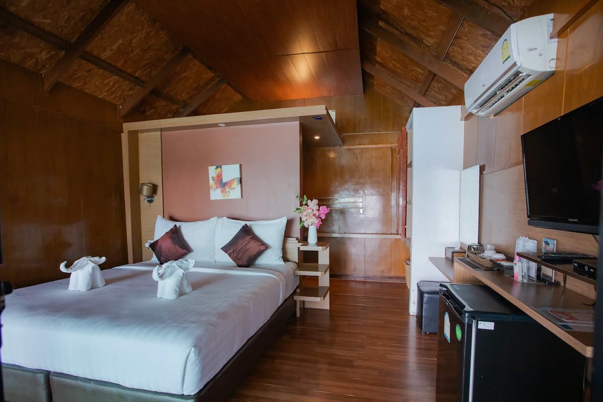 A hotel room with a wooden ceiling, a bed with white bedding, a small bedside table with a vase of flowers, a television, and a refrigerator.