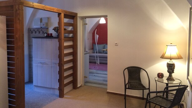 Een kamer met een houten ladder, een stoel, een tafel met een lamp, en een uitzicht op een rode muur en een bank op de achtergrond.