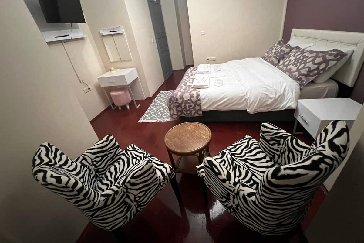 Un dormitorio con una cama, dos sillas de estampado de cebra, una mesita redonda pequeña, una mesita de noche blanca, un escritorio blanco con un taburete rosa, un televisor montado en la pared y una alfombra estampada.