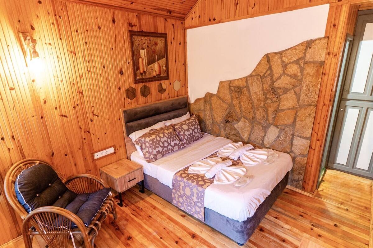 Una habitación con paneles de madera y una pared de acento de piedra, una cama con ropa de cama estampada, una mesita de noche, una silla y un cuadro en la pared.