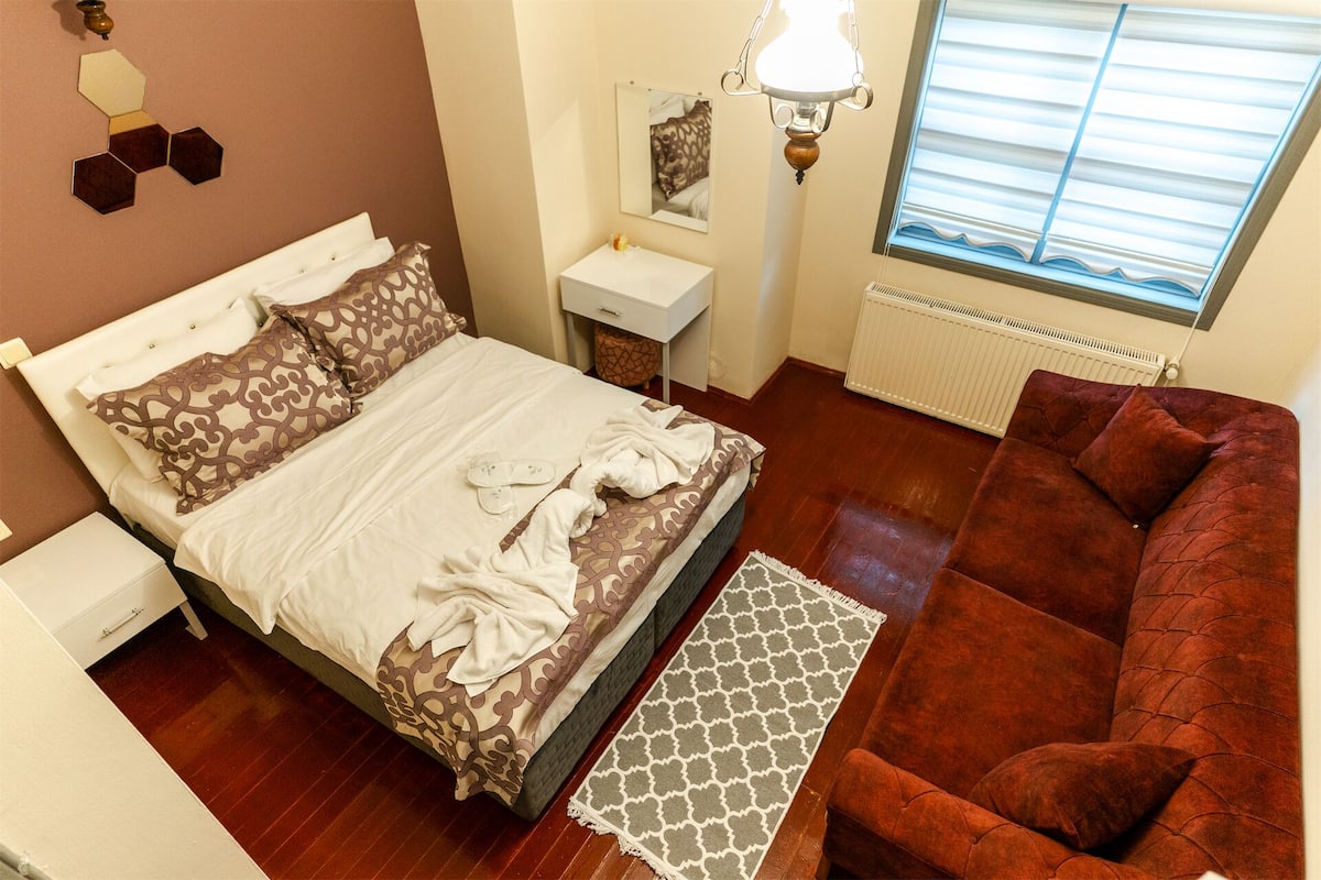 Un dormitorio con cama, mesita de noche, cómoda, sofá rojo, alfombra estampada, ventana con persianas e iluminación empotrada en la pared.
