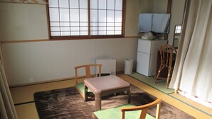 Una habitación japonesa tradicional con suelo de tatami, una mesa baja, dos sillas, un televisor y un refrigerador pequeño.
