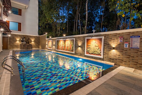 A swimming pool with a checkered tile pattern, surrounded by a brick wall with framed artwork and a sign indicating pool rules. - Crown Valley Thekkady (Peermade)