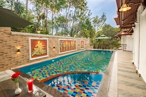 A swimming pool with a colorful mosaic floor, surrounded by a brick wall with framed artwork, and a table with a drink and a rose. - Crown Valley Thekkady (Peermade)