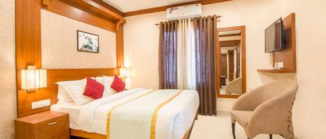 A hotel room with a wooden headboard, a bed with white linens and red pillows, a bedside table with a lamp, a chair, a flat-screen TV mounted on the wall, and a large mirror above the bed.
