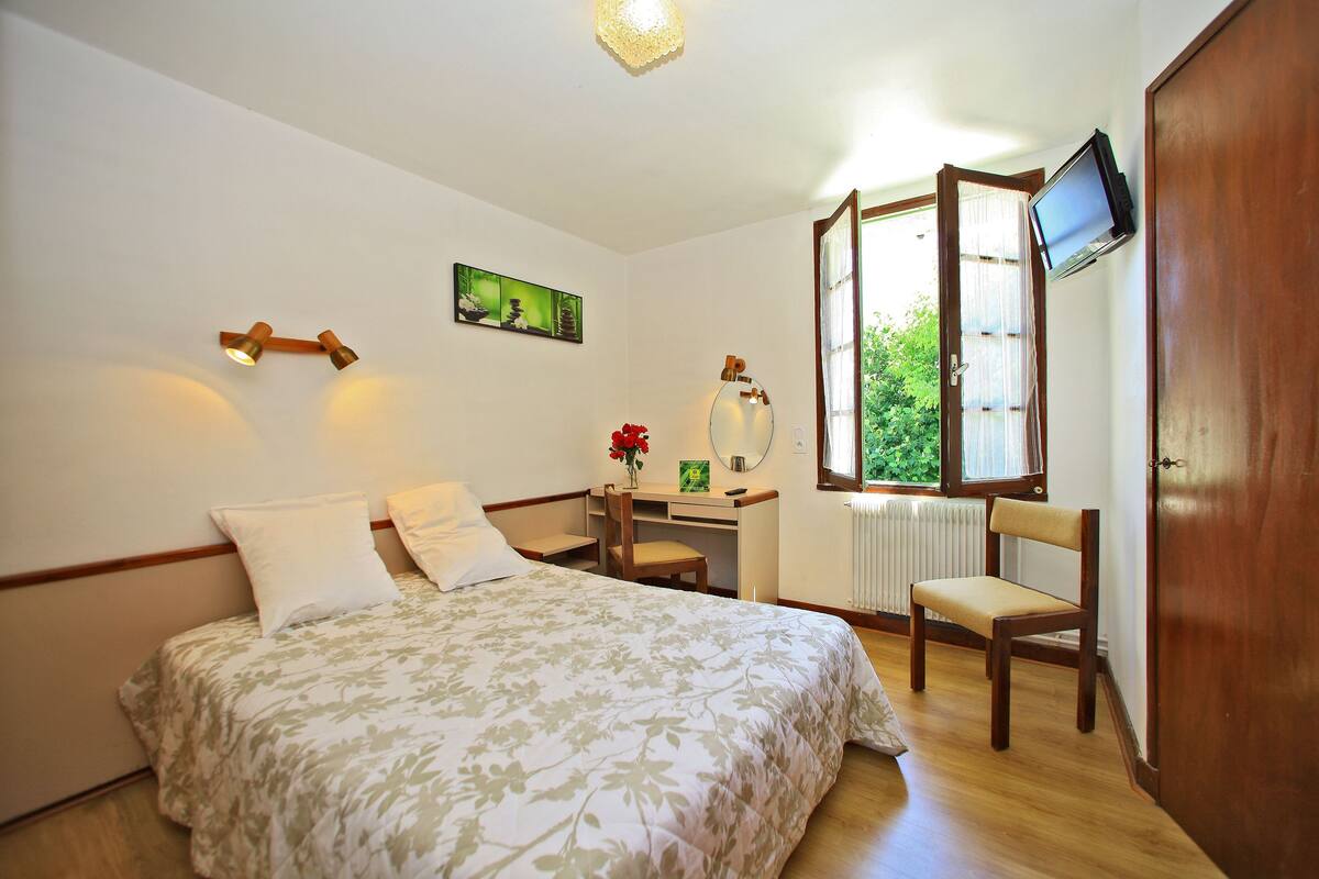 A bedroom with a bed, a desk, a chair, a TV, and a window.