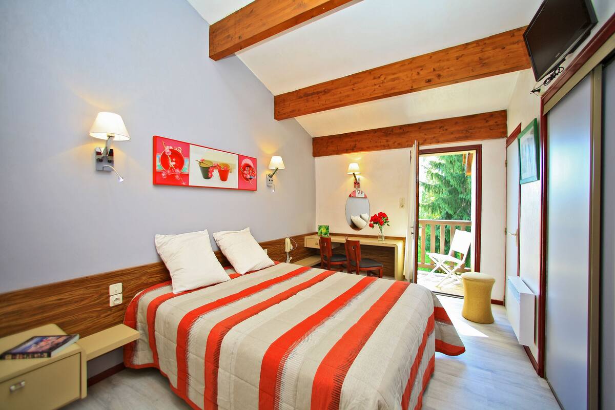 A bedroom with a bed, bedside table, lamp, artwork, and a view of the outdoors.