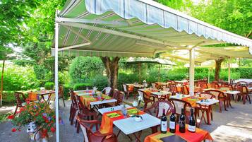 Un espace de restauration en plein air, avec des tables et des chaises disposées sous une tonnelle, le tout entouré de verdure.