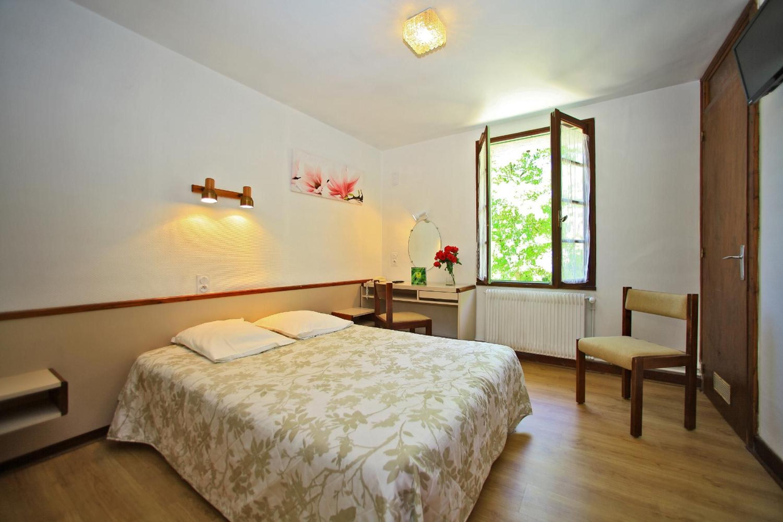 Quarto com cama, escrivaninha, cadeira, janela com vista para área verde e luminária na parede.