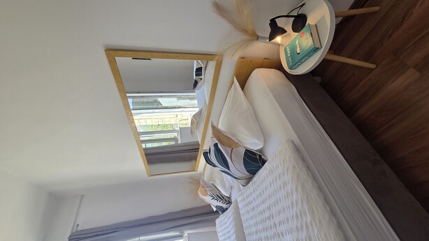 Un dormitorio con cama, una mesita de noche con un libro y audífonos, una ventana con vistas, y un espejo en la pared.