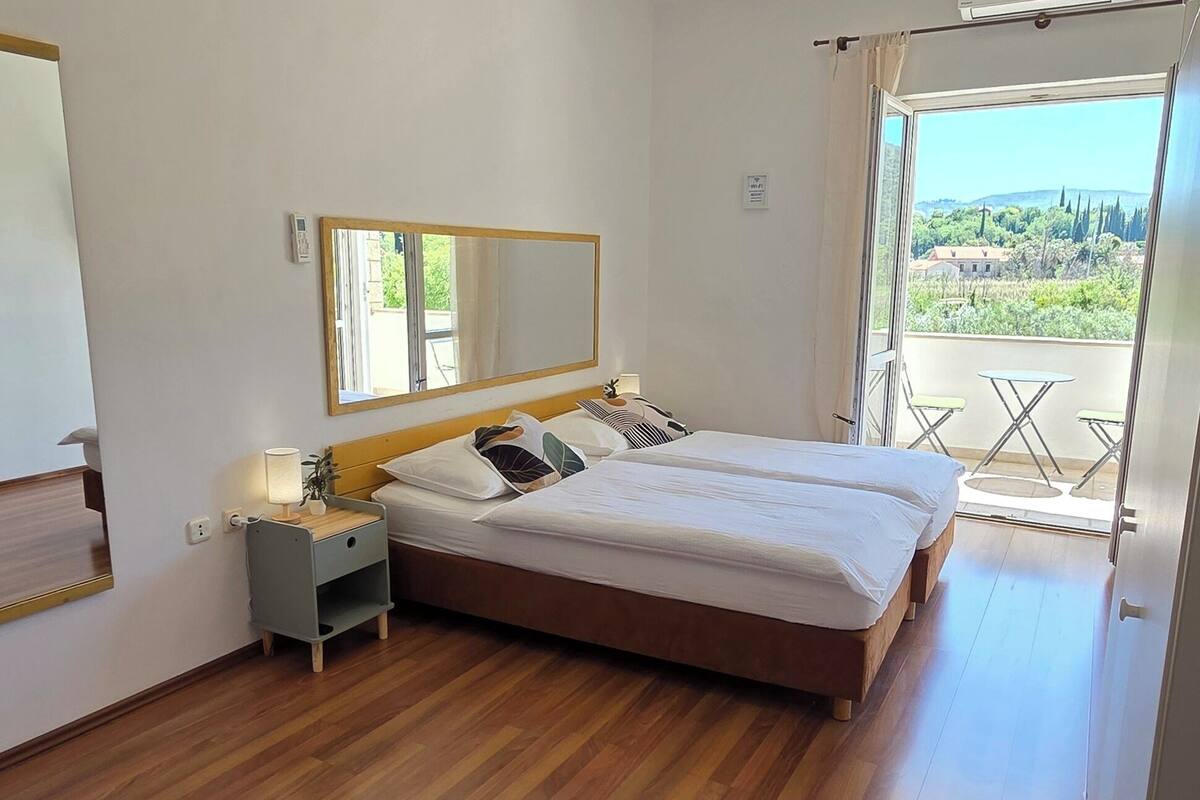 A bedroom with a large bed, a bedside table with a lamp, a mirror, and a view of the outdoors through a large window.