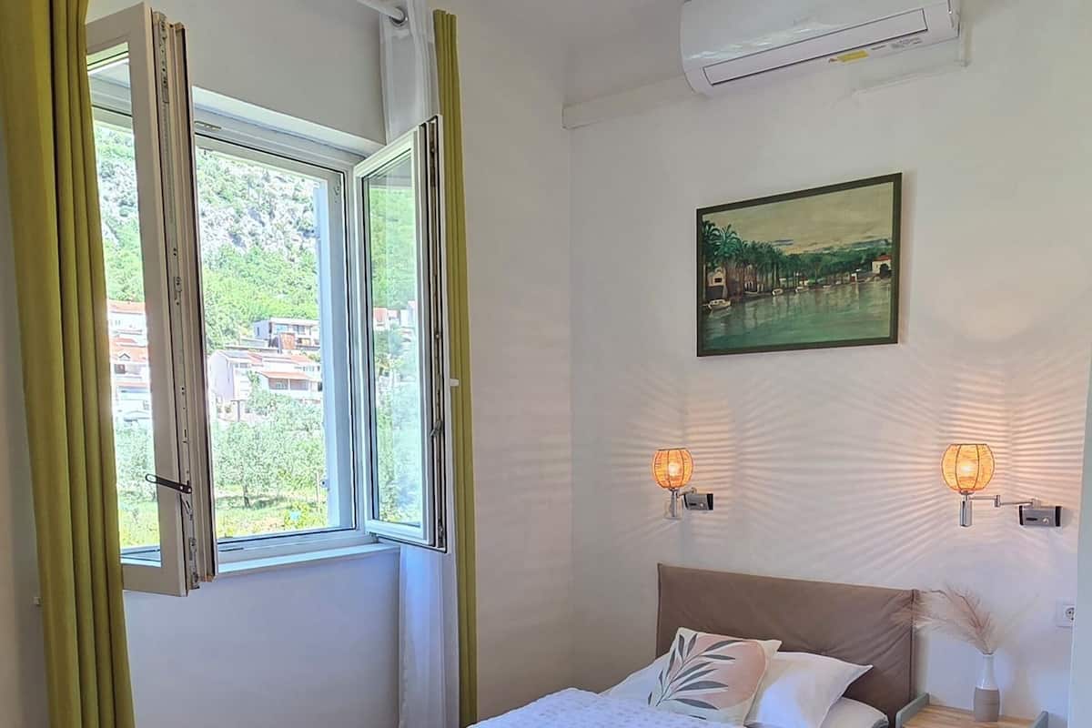 A bedroom with a bed, an air conditioner, a window with curtains, a wall-mounted picture, and bedside lamps.