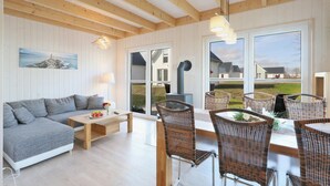 A cozy living room with a sofa, dining table, wicker chairs, and a large window offering a view of the outdoors. - Lovely home in OstseeResort Olpenitz with sauna (OstseeResort Olpenitz)
