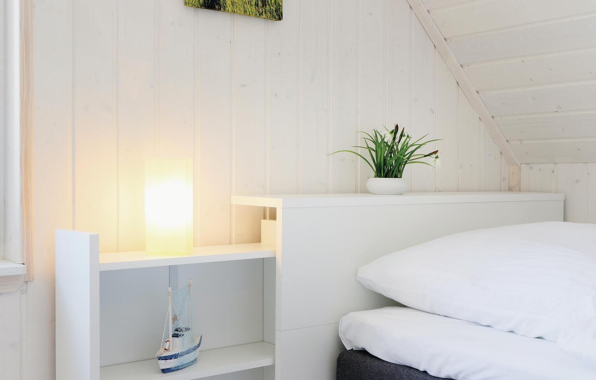 A bedroom with a white bed, a small shelf with a model ship, a potted plant, and a white wall.