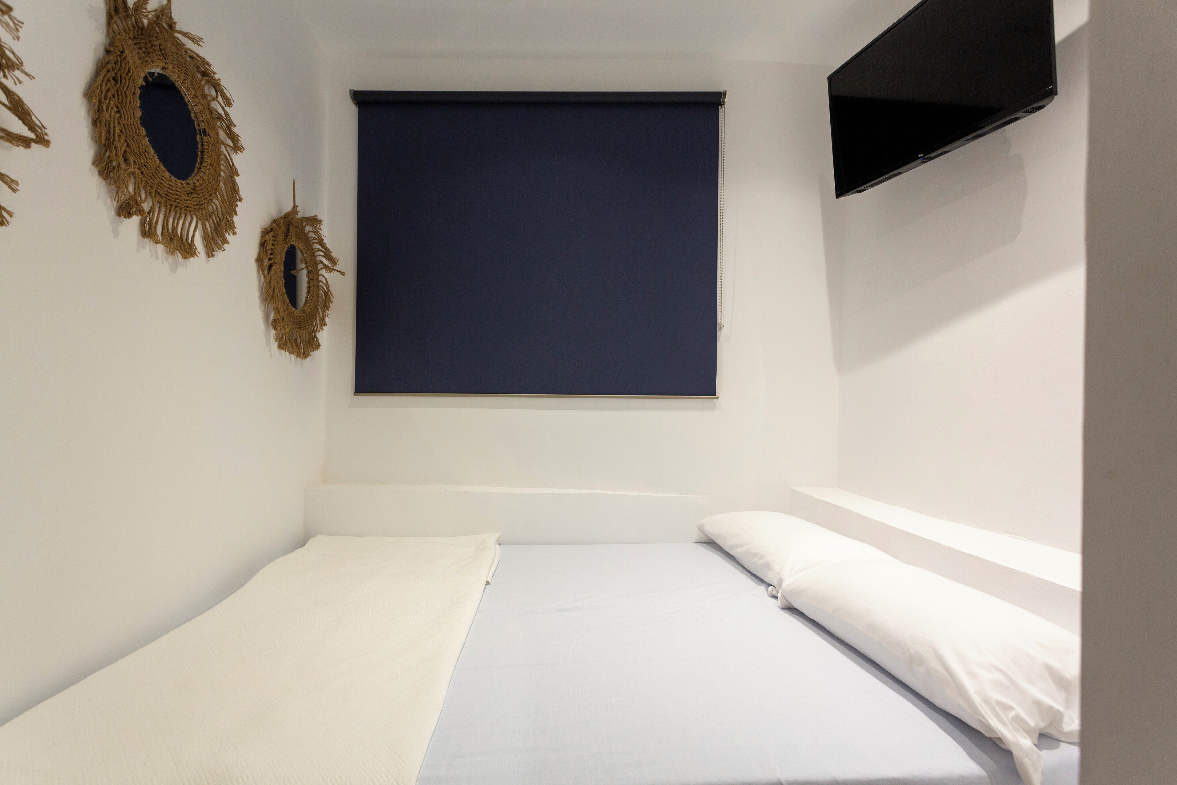 Una habitación pequeña con cama individual, persiana azul oscuro, televisor montado en la pared y dos adornos de macramé colgados en la pared.