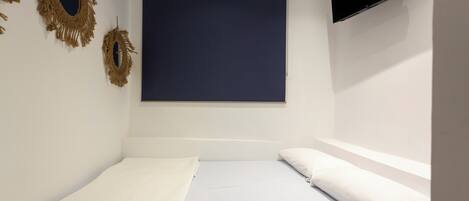Una habitación pequeña con cama individual, persiana azul oscuro, televisor montado en la pared y dos adornos de macramé colgados en la pared.