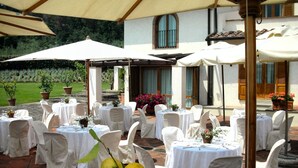 An outdoor patio with round tables covered with white tablecloths and white chairs, set up under large umbrellas. - Tenuta le Viiste (Scandicci)