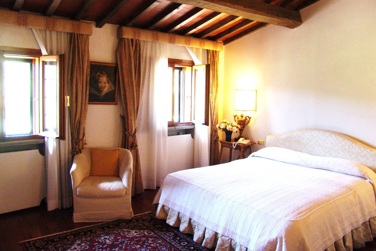 A bedroom with a bed, a chair, a nightstand with a lamp, and a window with curtains.
