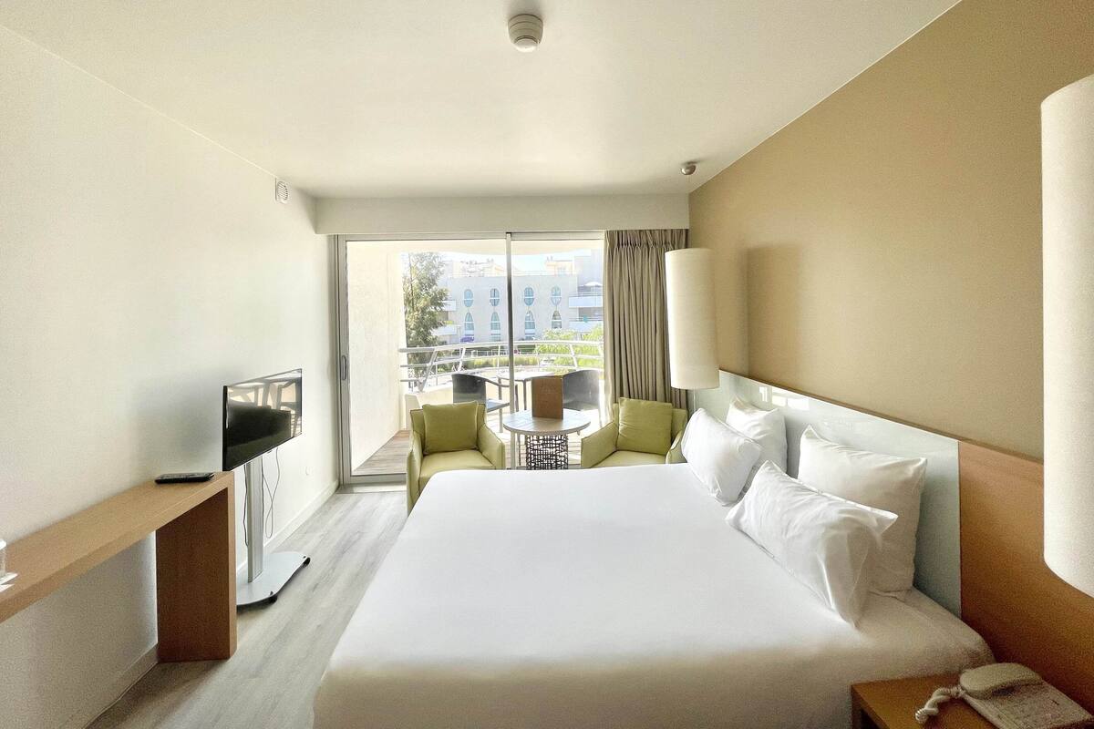 Een hotelkamer met een groot bed, een bureau met een computer, een stoel en een kleine tafel met een lamp.