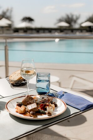 Una mesa de comedor con un plato de comida, un vaso de agua y un vaso de vino blanco. La mesa está al aire libre, con una piscina al fondo.