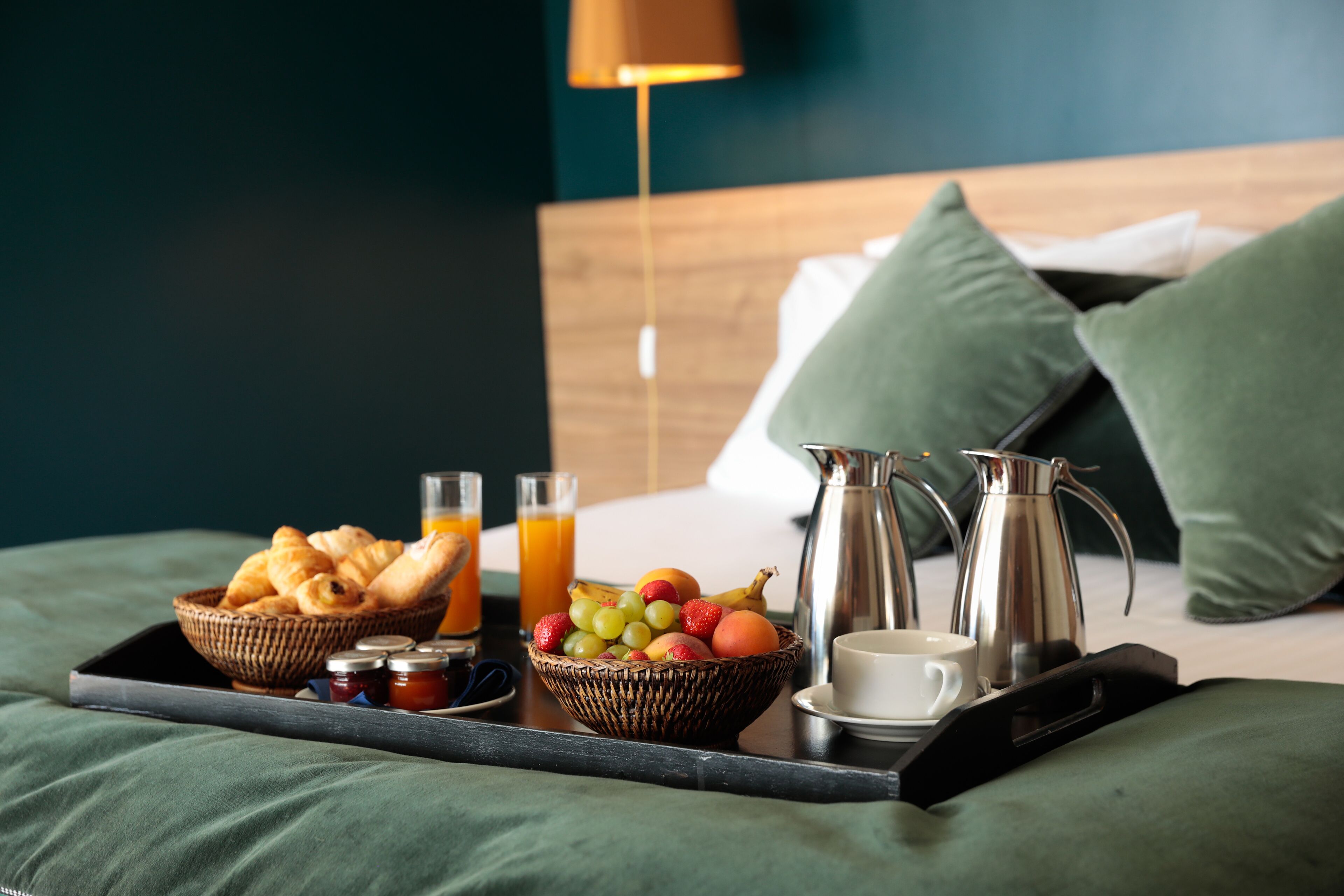 Frühstückswagen mit Croissants, Obst und Getränken auf einem Bett mit grünen Kissen und einem Holzkopfende.