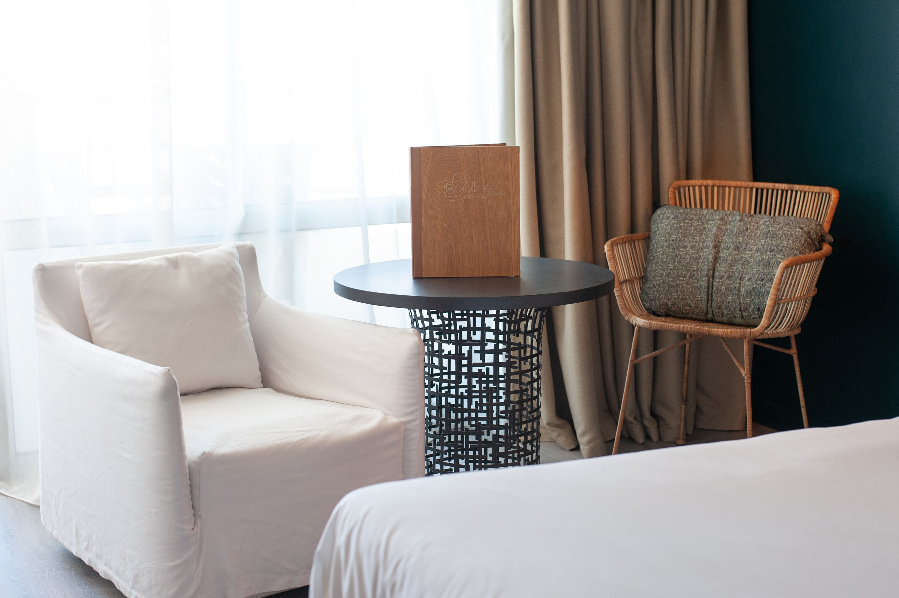 Une chambre d’hôtel avec un lit, un fauteuil blanc, une chaise en osier et une petite table ronde sur laquelle se trouve une boîte en bois.