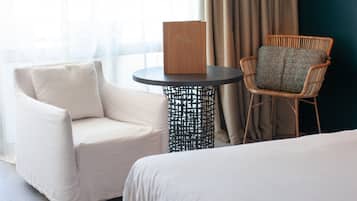 Une chambre d’hôtel avec un lit, un fauteuil blanc, une chaise en osier et une petite table ronde sur laquelle se trouve une boîte en bois.