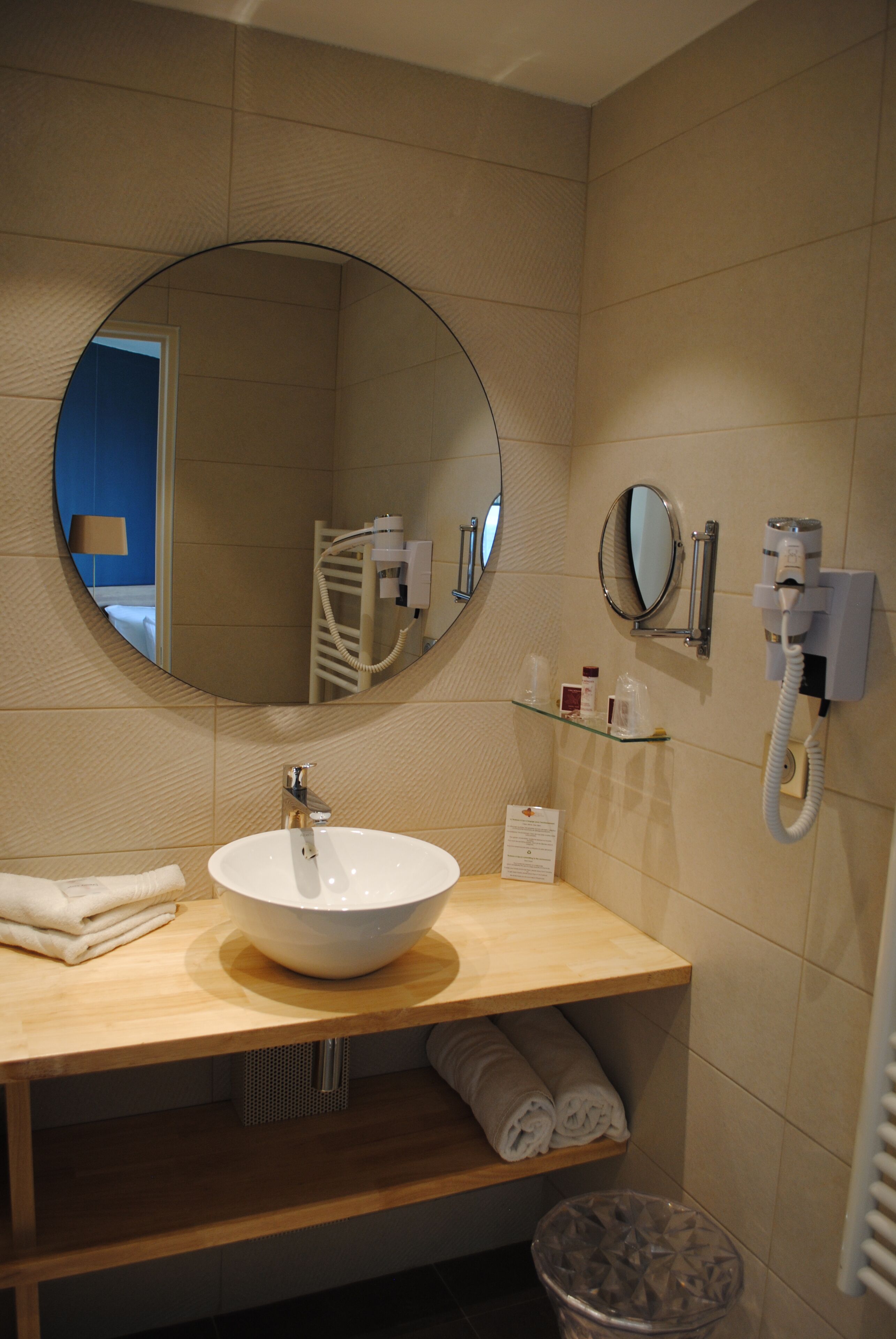Une salle de bain avec un miroir rond, un lavabo, un porte-serviettes, un sèche-cheveux et une poubelle.