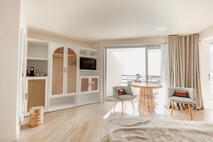 Una habitación de hotel moderna con un ventanal grande, una mesa redonda de comedor, dos sillones, un armario con televisión y vistas al balcón.