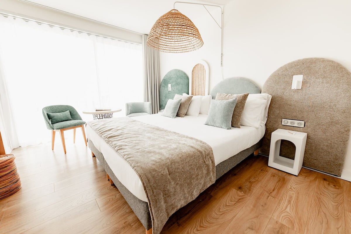 Een moderne slaapkamer met een groot bed, een klein nachtkastje, een stoel, een geweven lamp en een wandcontactdoos.